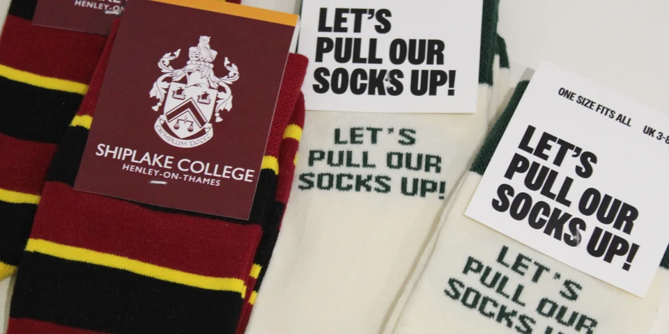 Maroon, black and gold striped socks beside cream socks reading LET'S PULL OUR SOCKS UP, with Shiplake College, Henley-on-Thames swing tags laid on top.