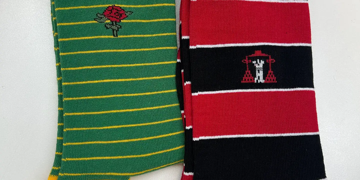 Two socks on a white surface: a green and gold pair with a red rose crest, and a red and black hooped pair with a tower crest.
